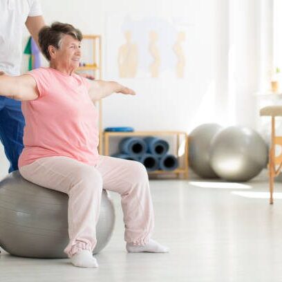 Elderly senior ward trying to maintain balance while sitting on a grey fit ball while being supported by physiatrist